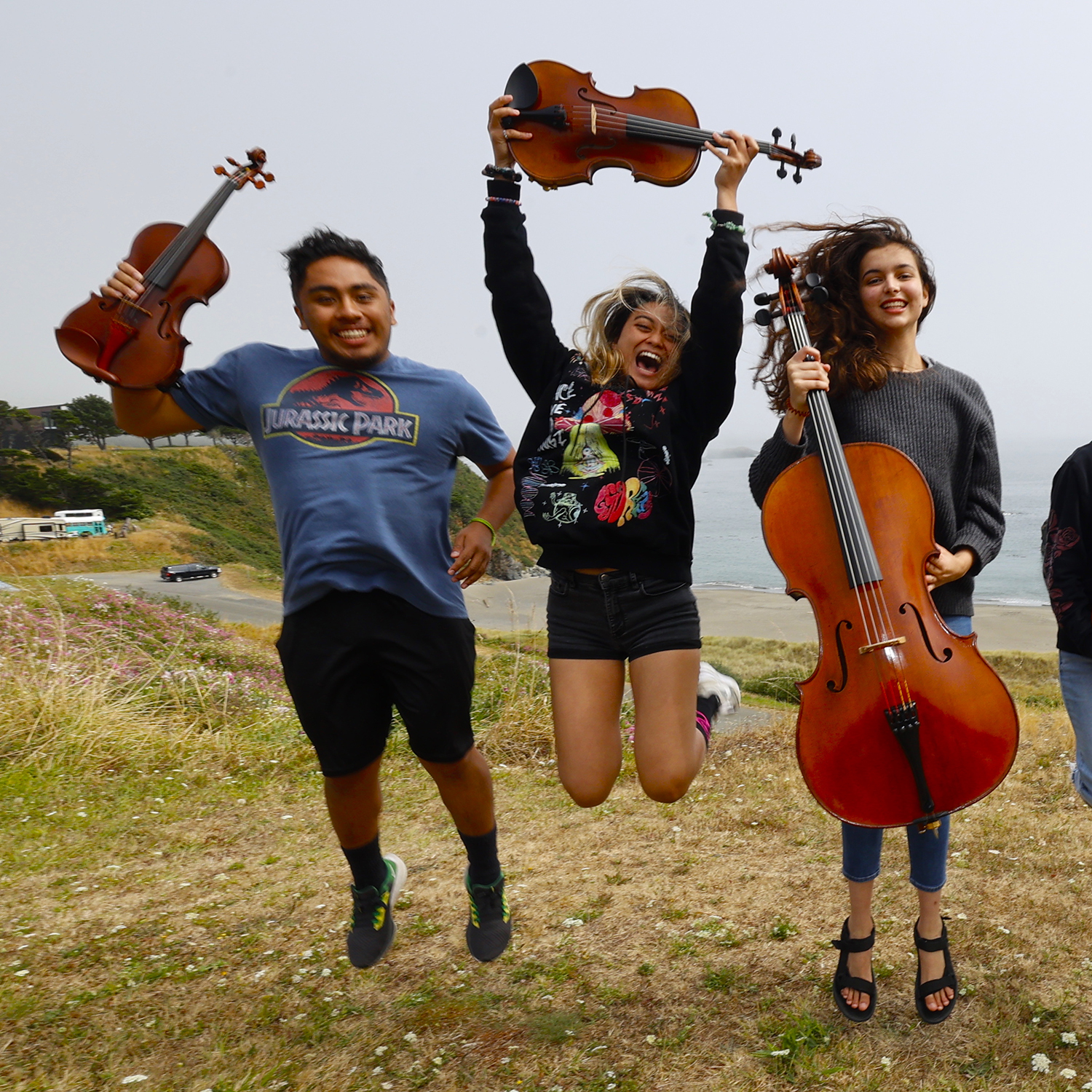 Three string young musicians jump in delight outside with their instruments.
