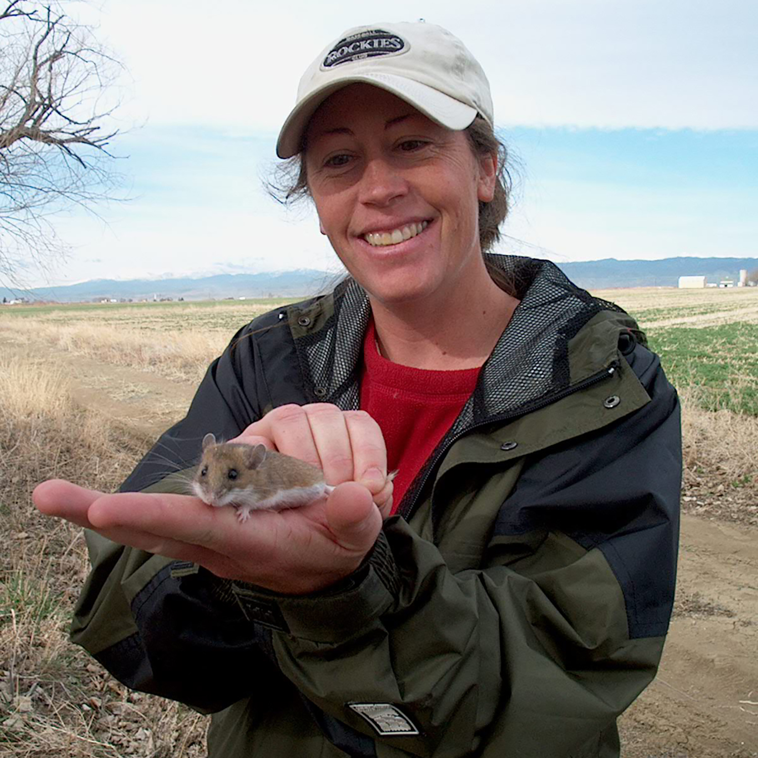 a woman standing outside smiles with an outstretched hand where she is holding a small mouse